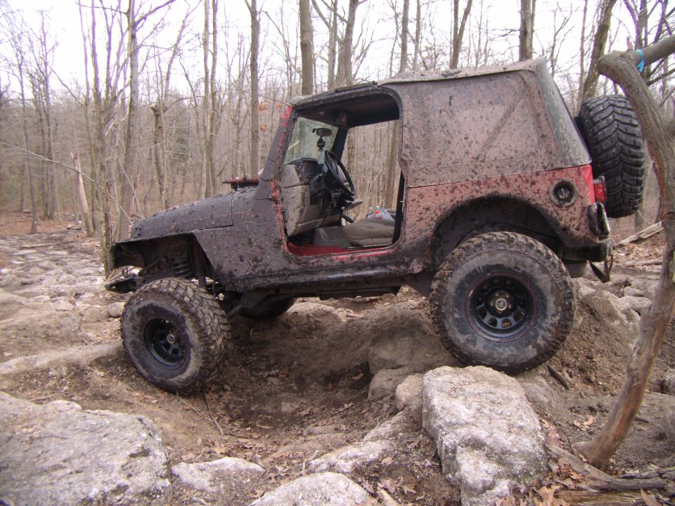 Muddy Jeep Wrangler navigating rocks on a wooded trail. The 2003 Jeep Wrangler is covered in mud