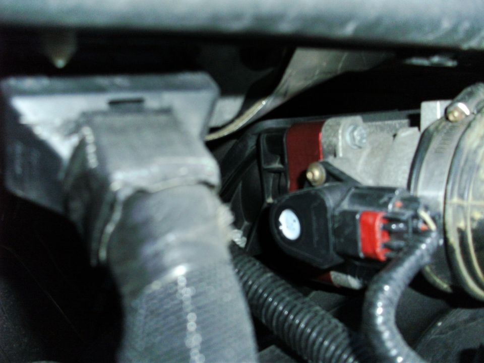 Close-up of an engine sensor with red and black connectors, attached to wiring and hoses