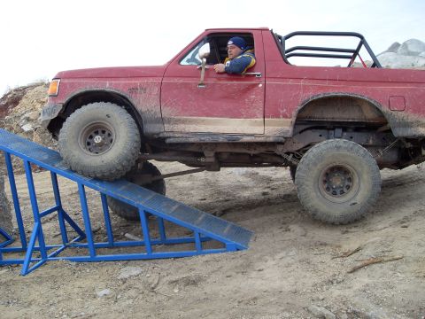 1987 Ford Bronco - A red SUV with off-road modifications navigates a blue ramp during an off-road event.