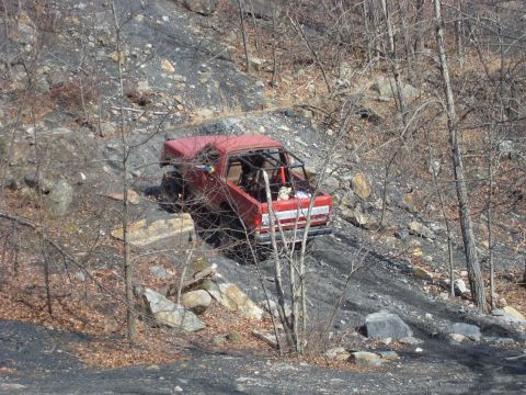1987 Ford Bronco - A red truck navigates a steep, rocky incline in a wooded area.