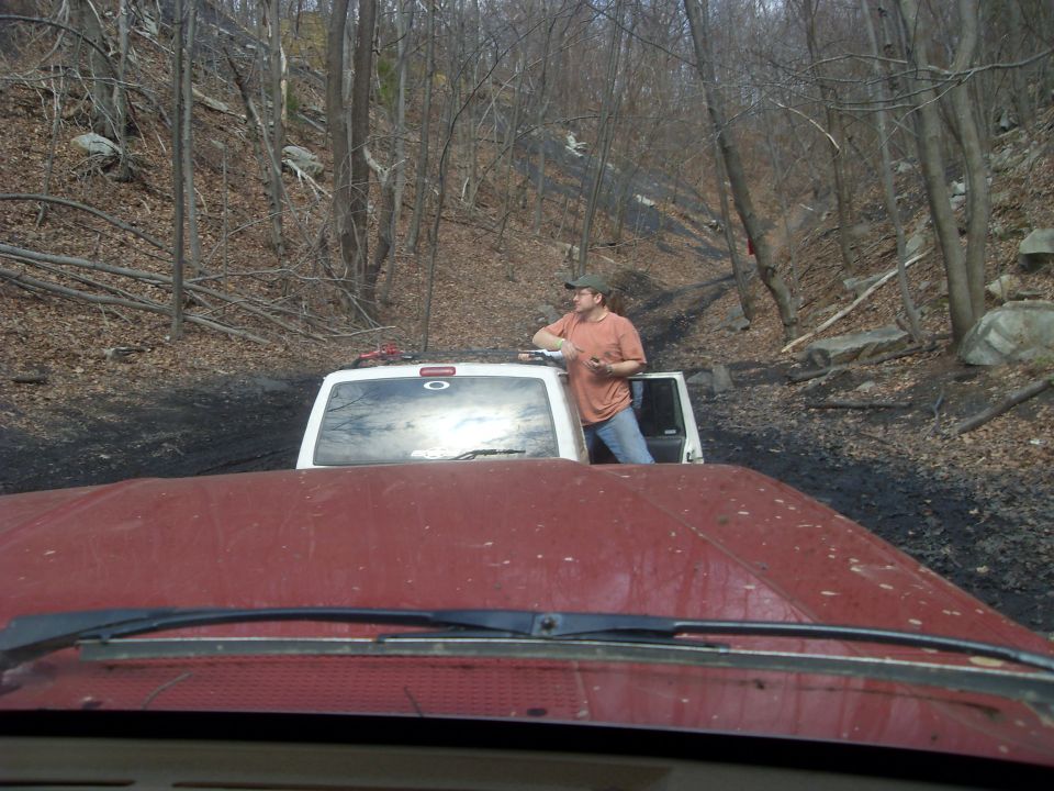 View from inside a red vehicle looking at a white SUV on a muddy off-road trail with trees and hills in the background