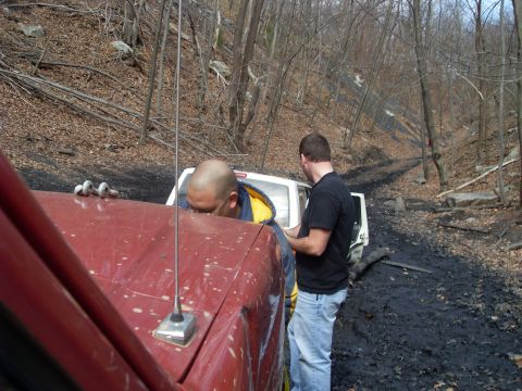 1987 Ford Bronco - Two men assess a white SUV stuck in a muddy, wooded trail, with a red truck in the foreground.