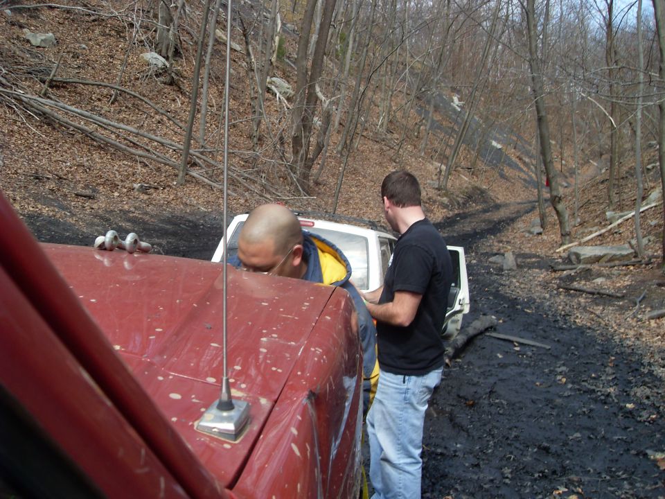 Red truck hood with antenna, white SUV stuck in mud, two men looking at it