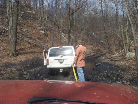 1987 Ford Bronco - Two people attempt to free a white Jeep Cherokee stuck in thick mud on a wooded trail.