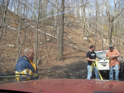 1987 Ford Bronco - People are preparing to tow a white SUV in a wooded, off-road environment.