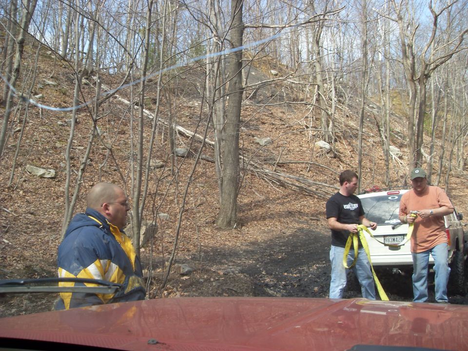 Two guys with a tow strap by a white SUV in a wooded area. Another person in a blue and yellow jacket is visible