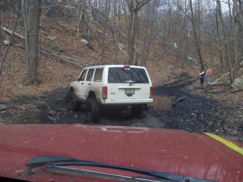 1987 Ford Bronco - A white Jeep Cherokee navigates a muddy trail in a wooded area.