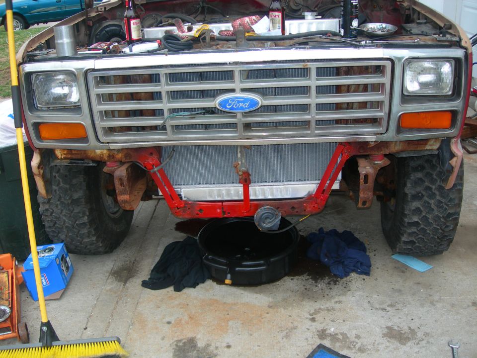 Front view of a rusty red Ford Bronco with hood open, radiator exposed, and maintenance supplies on the ground.