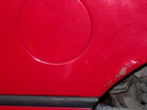 1984 Ford Bronco - A close-up view of the lower rear quarter panel of a red vehicle, showing a circular fuel filler door and some rust spots near the bottom e…