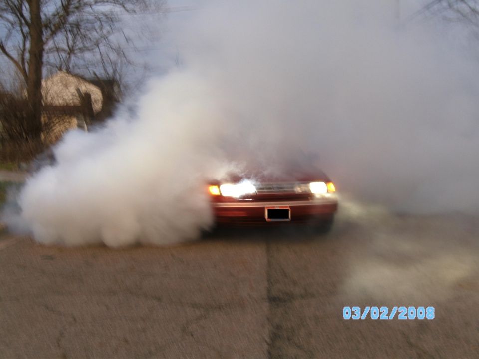 Dark red sedan doing a burnout, headlights on, mostly hidden by thick white tire smoke on a street