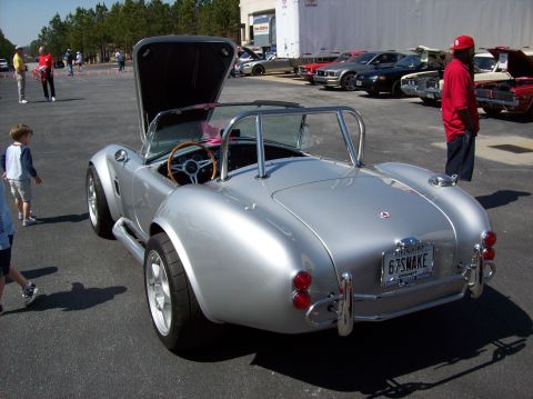 2007 Ford Mustang - A silver AC Cobra replica with its hood open is parked on asphalt. Several other cars are visible in the background, along with people.