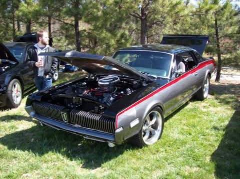 2007 Ford Mustang - A black and silver classic coupe with a red stripe is parked on a grassy lawn with its hood open, revealing the engine. A man stands to the…