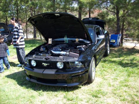 2007 Ford Mustang - A black Ford Mustang with its hood open is parked on a grassy area with trees in the background. A young boy and an adult are standing near…