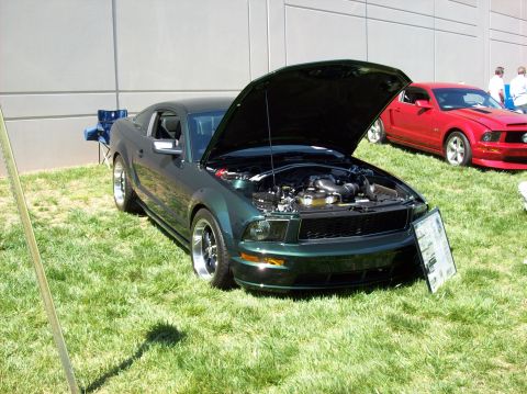 2007 Ford Mustang - A dark green 2007 Ford Mustang coupe with its hood open is parked on a grassy lawn in front of a building. A red Ford Mustang is parked beh…