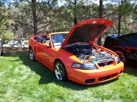 2007 Ford Mustang - An orange convertible Ford Mustang is parked on a grassy area with its hood open, revealing the engine. A black car is visible behind it.