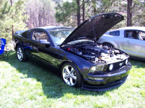 2007 Ford Mustang - A black Ford Mustang GT with its hood open is parked on a grassy lawn in front of trees. Another silver car is visible behind it.