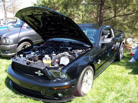 2007 Ford Mustang - A black Ford Mustang with its hood open is parked on a grassy area, showcasing its engine. Another vehicle, a gray pickup truck, is visible…