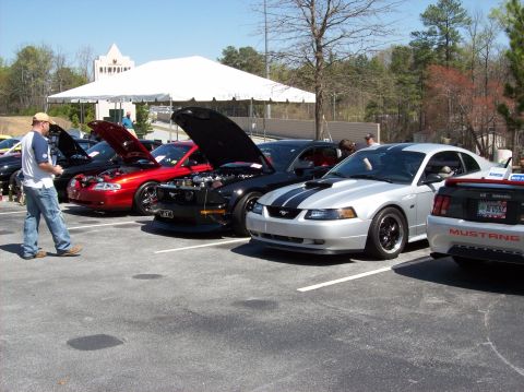 2007 Ford Mustang - A line of Ford Mustang cars are parked in a lot with their hoods open, showcasing their engines. A man in a cap and jeans walks past the ca…