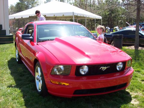2007 Ford Mustang - A bright red Ford Mustang is parked on a grassy lawn in front of a white tent. A young girl in a pink shirt is looking at the car.