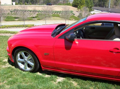 2007 Ford Mustang - Side profile of a red Ford Mustang GT with a chrome wheel and a GT emblem on the fender. The car is parked on a grassy area with a landscap…