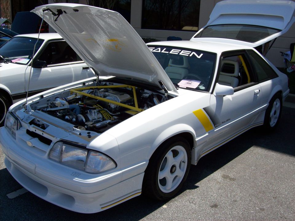 White Saleen Mustang with hood and trunk open, showing engine bay with yellow roll cage.