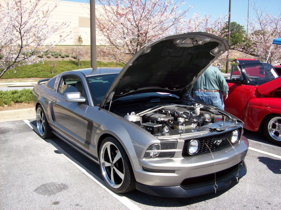 Silver Ford Mustang with hood open, showing engine. Black and silver wheels. Parked next to a red pickup truck