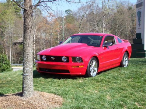 2007 Ford Mustang - Red Ford Mustang with a hood scoop parked on a grassy area near a tree and building.