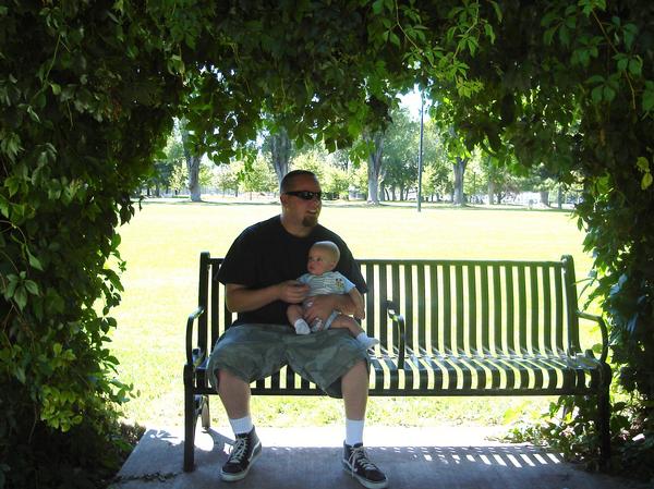 Man holding a baby while sitting on a park bench under leafy trees. A watermark reads SUPERMOTORS