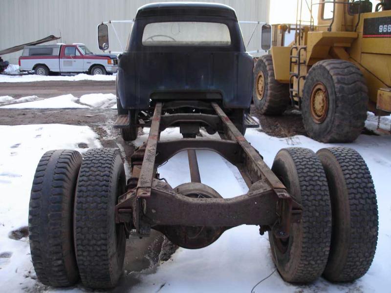 Cab-and-chassis vintage Ford truck from the rear, bare frame and dual rear wheels, next to a yellow loader