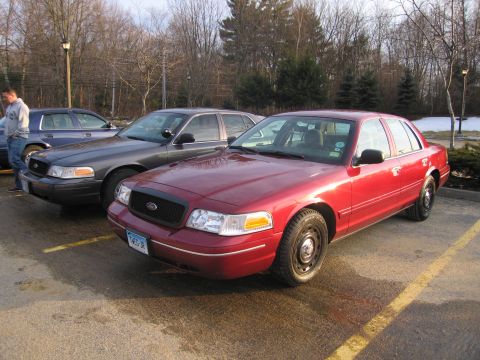2001 Ford Crown Victoria - Two Ford Crown Victorias are parked in a lot, one red and one dark gray, with a person standing near the gray one.