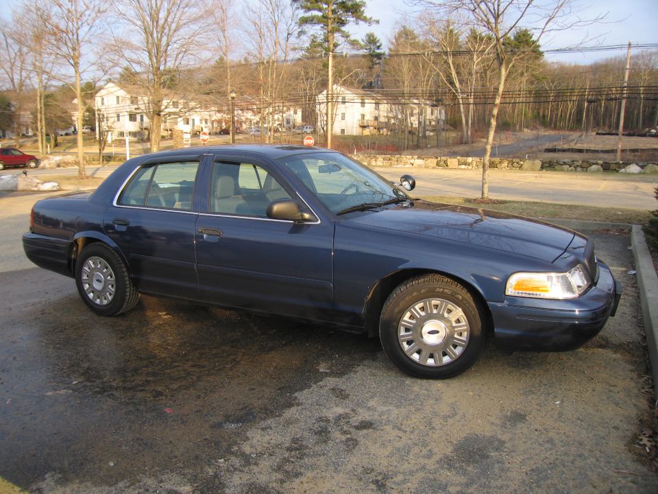 Dark blue Ford Crown Victoria parked on wet asphalt. It has a police-style spotlight on the driver's side