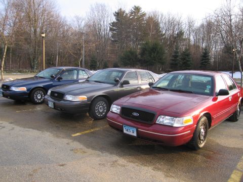 2001 Ford Crown Victoria - Three Ford Crown Victorias are parked in a lot. The closest car is red, and the other two are dark colors.
