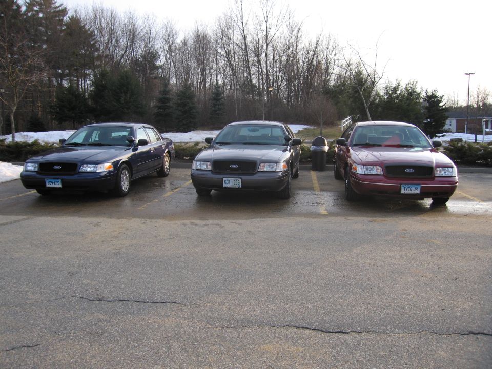 Three Ford Crown Victorias parked in a lot. The leftmost is dark blue, the middle is dark gray, and the rightmost is maroon. All are facing forward