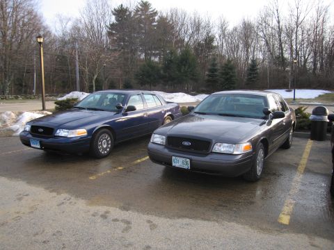 2001 Ford Crown Victoria - Two Ford Crown Victorias, a blue one and a gray one, are parked side-by-side in a lot on a cloudy day.