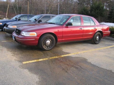 2001 Ford Crown Victoria - Three Ford Crown Victorias are parked in a lot, one in red and two in darker colors.