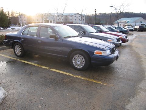 2001 Ford Crown Victoria - A line of dark blue Ford Crown Victorias are parked in a lot with the sun setting in the background.