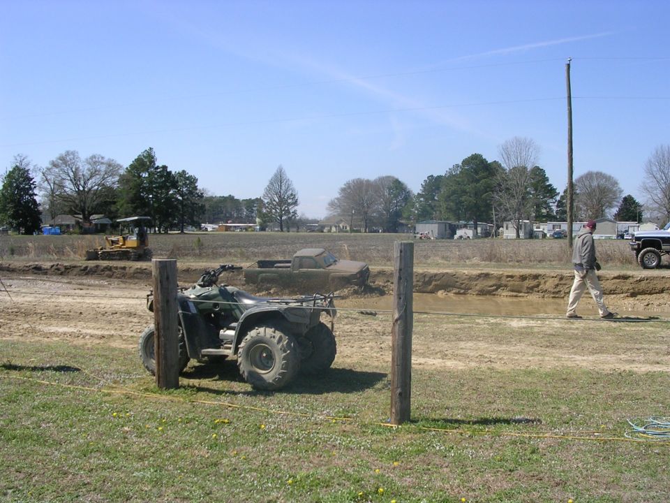 Green ATV in foreground, muddy ditch with pickup truck behind it. Person walking in background