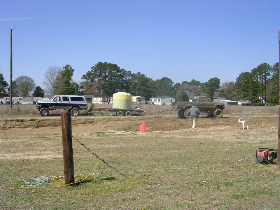 lifted white SUV towing a yellow tank trailer, and a green pickup truck in a muddy area