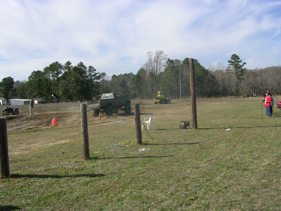 Lifted pickup truck on a dirt track, with an ATV and a yellow tractor nearby. People are watching