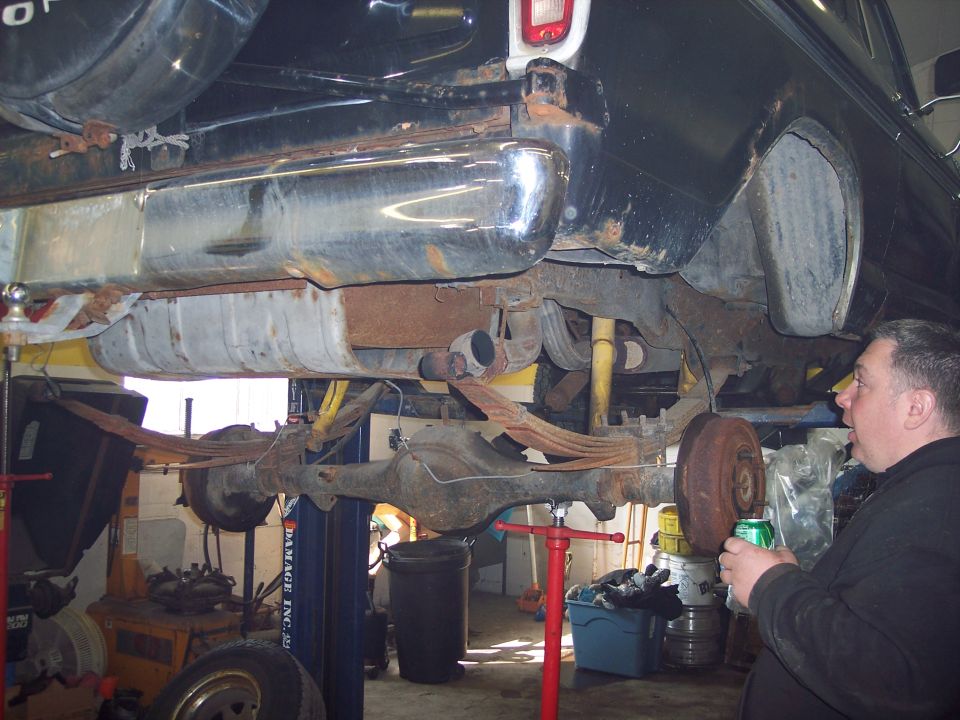 Underside of a vintage vehicle on a lift, showing fuel tank, leaf springs, and rear axle with rust.