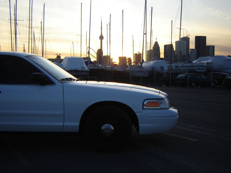 White Ford Crown Victoria side view with sailboats and city skyline at sunset.