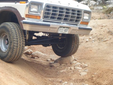 1979 Ford Bronco - The front passenger side of a white 1979 Ford Bronco is shown at an angle, with the front tire resting on a rock on a dirt road. The vehicl…