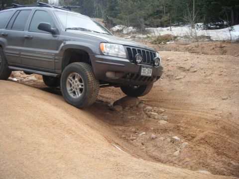 1979 Ford Bronco - A dark gray Jeep Grand Cherokee is navigating an off-road trail with its front right tire on a rocky incline. The vehicle is angled towards…