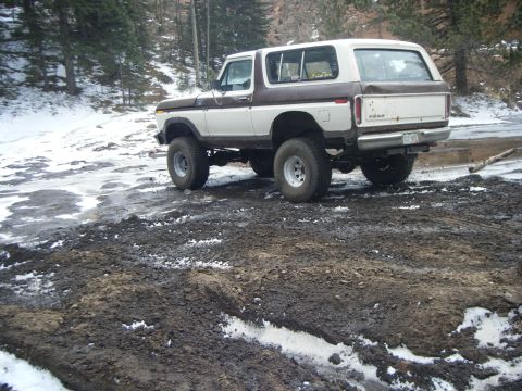 1979 Ford Bronco - A two-tone brown and white Ford Bronco is parked on a muddy and snowy off-road trail. The vehicle is viewed from the rear-three-quarter ang…