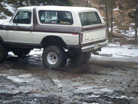 1979 Ford Bronco - The right rear side of a white and brown 1979 Ford Bronco is shown driving through a muddy and snowy area. The vehicle has large off-road t…
