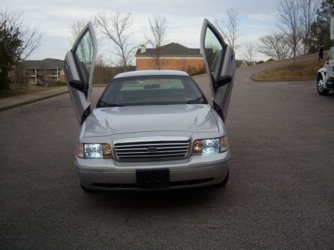 1993 Ford Crown Victoria - A silver Ford Crown Victoria with its doors open upwards, parked on an asphalt surface. The car has custom headlights and a chrome grille,…