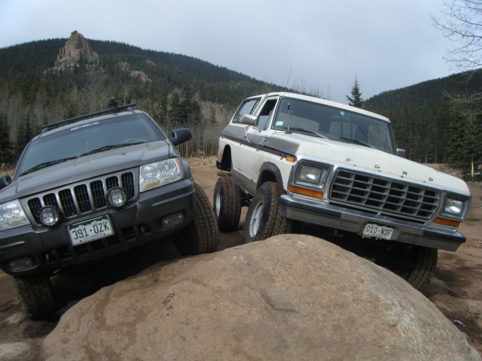 Gray Jeep Grand Cherokee and white Ford Bronco on a rock in a forest.