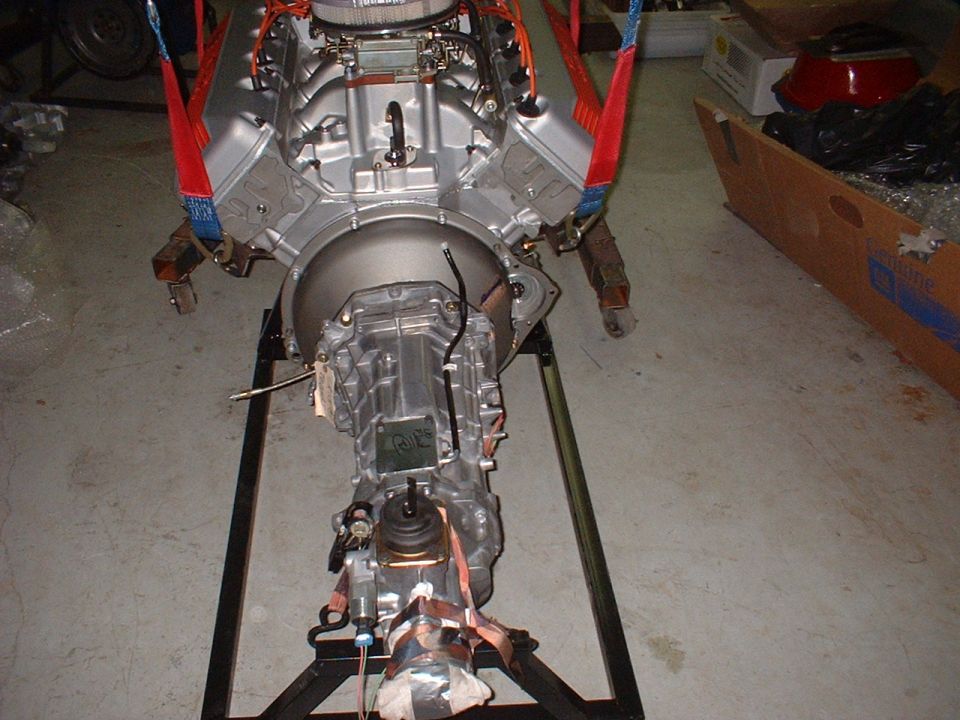 Silver V8 engine and transmission assembly on a stand, suspended by straps.