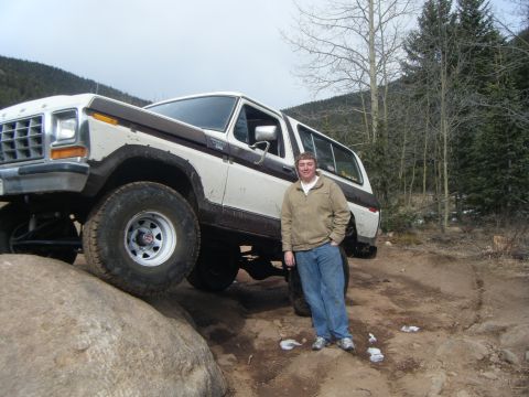 1979 Ford Bronco - A man stands next to a lifted, two-tone white and brown Ford Bronco on a rocky, wooded trail. The Bronco is angled with its front left whee…