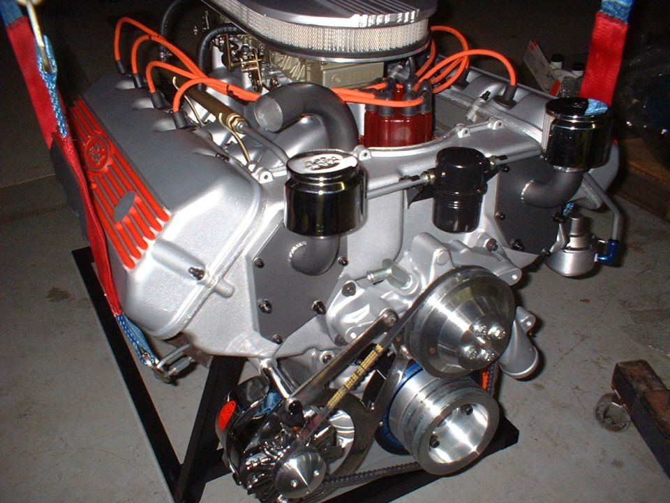 Silver V8 engine with orange wires and red valve cover accents on a black stand.
